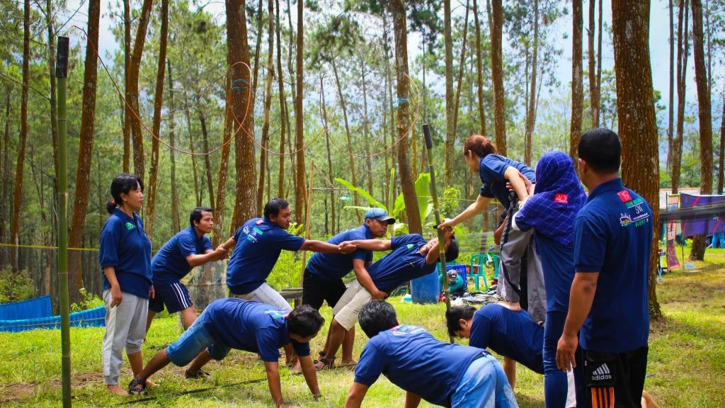 Peserta korporat berkaus seragam menyelesaikan tantangan team dynamics di ground hutan pinus bersama fasilitator First Outbound