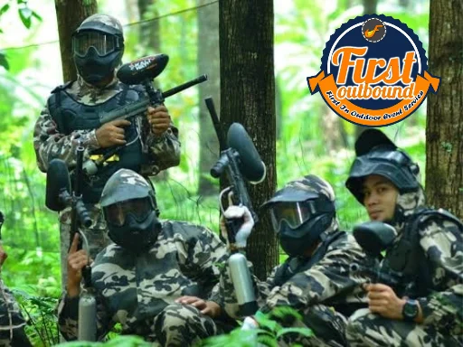 Regu paintball berseragam loreng, bermasker, dan membawa marker sedang berlindung di balik pohon saat war games hutan